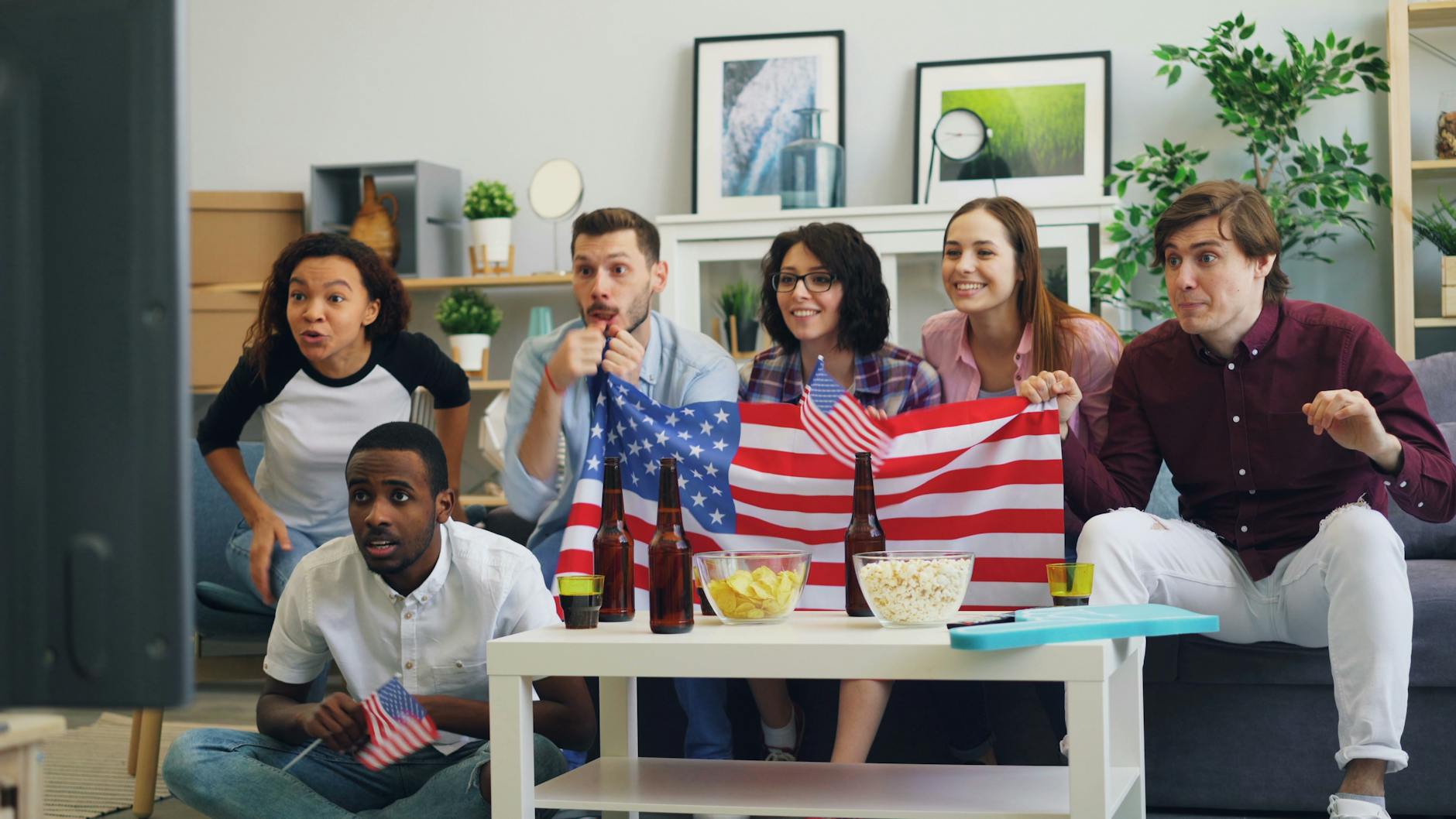 A lively group of friends celebrates while watching sports indoors, holding American flags and enjoying snacks.