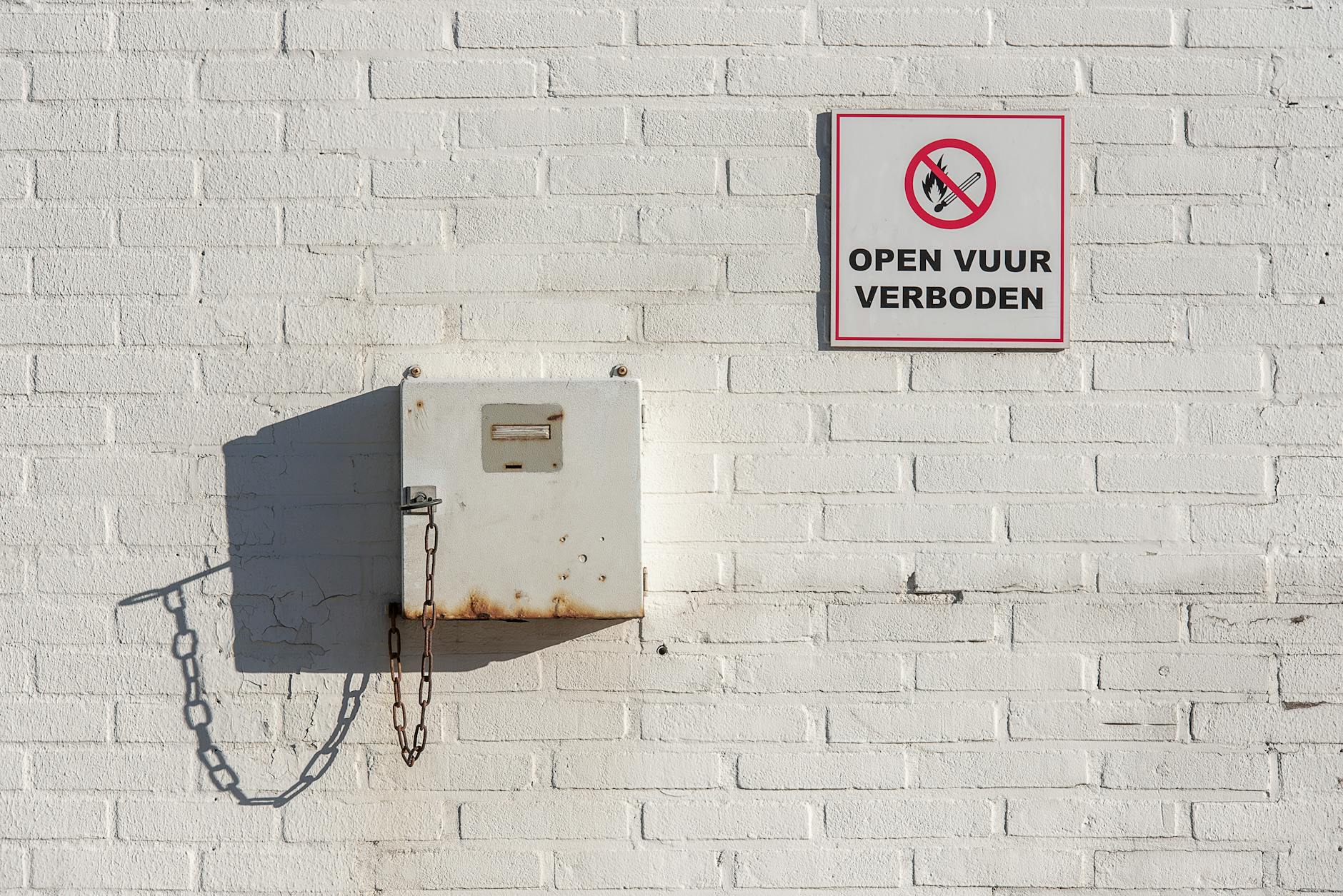 White wall with a no open fire sign and metal box with chain, casting shadow.