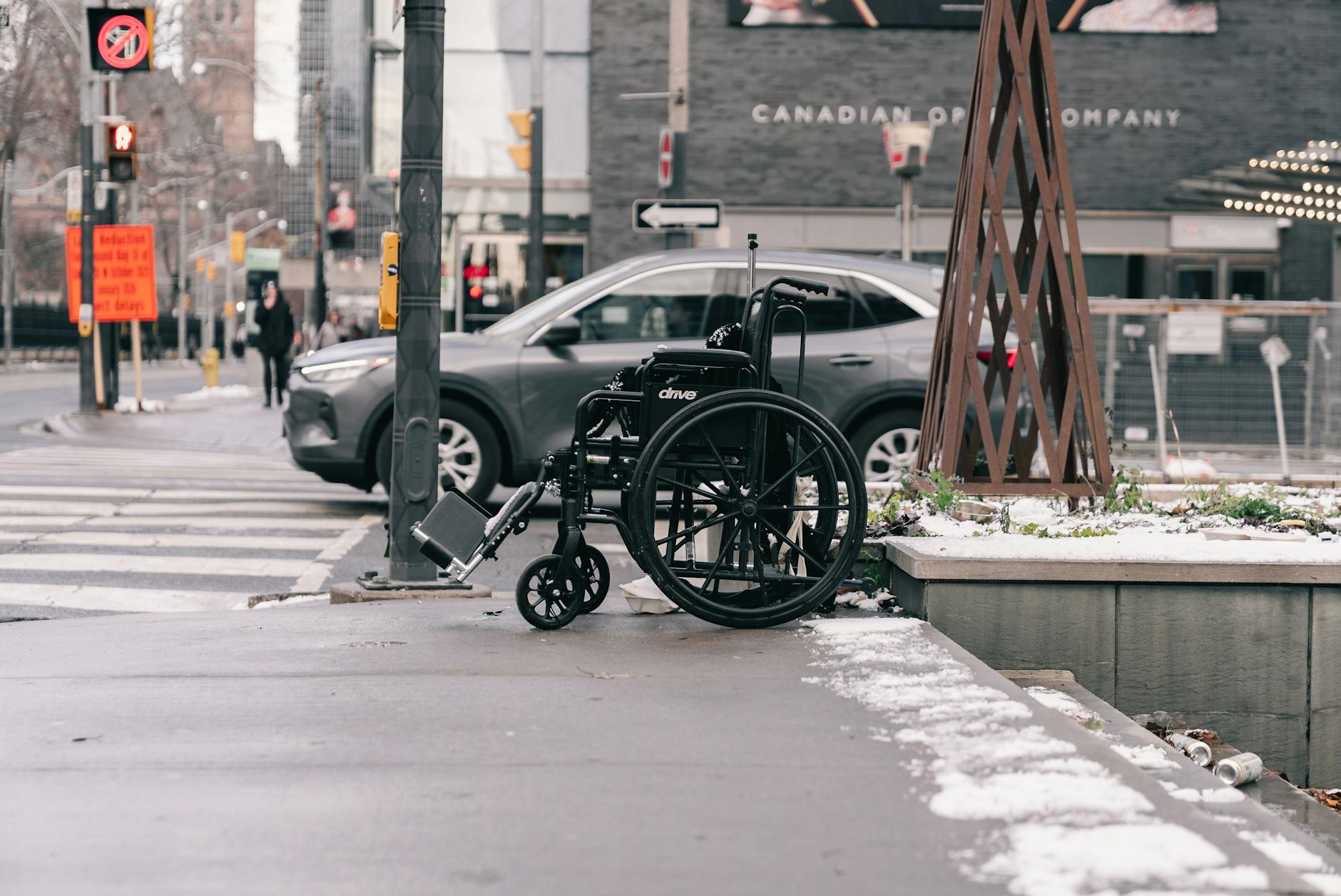 A wheelchair is parked on a snowy city sidewalk during winter with a car passing by.