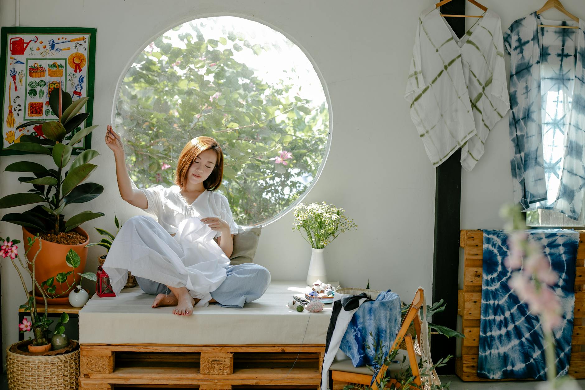 Full body of ethnic barefoot young lady in casual outfit sitting on seat at home while sewing white fabric near window and decorative elements