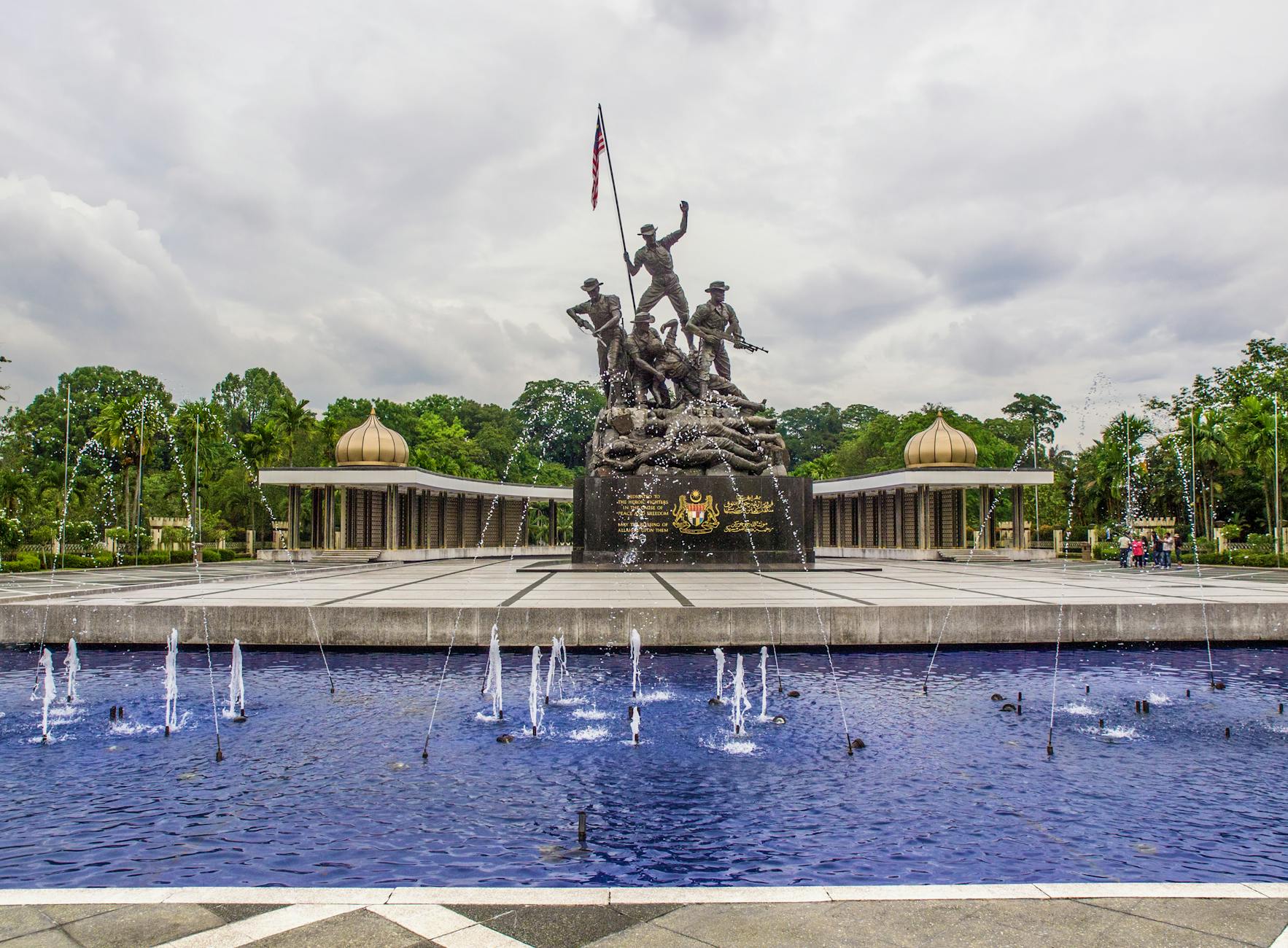 View of the National Monument in Kuala Lumpur, Malaysia, featuring a fountain in the foreground and dramatic clouds.