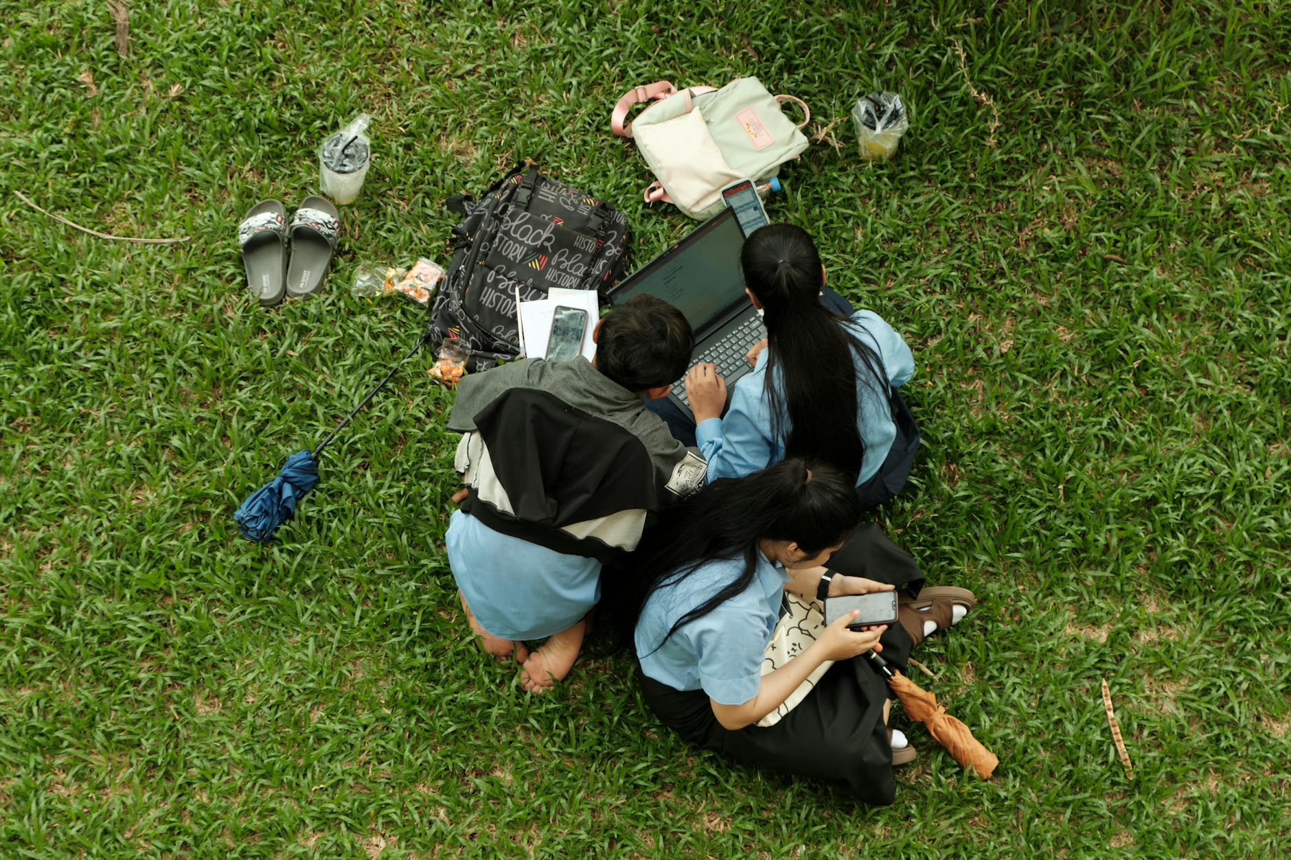 Top view of three friends using a laptop and phones on grass, enjoying a picnic outdoors.