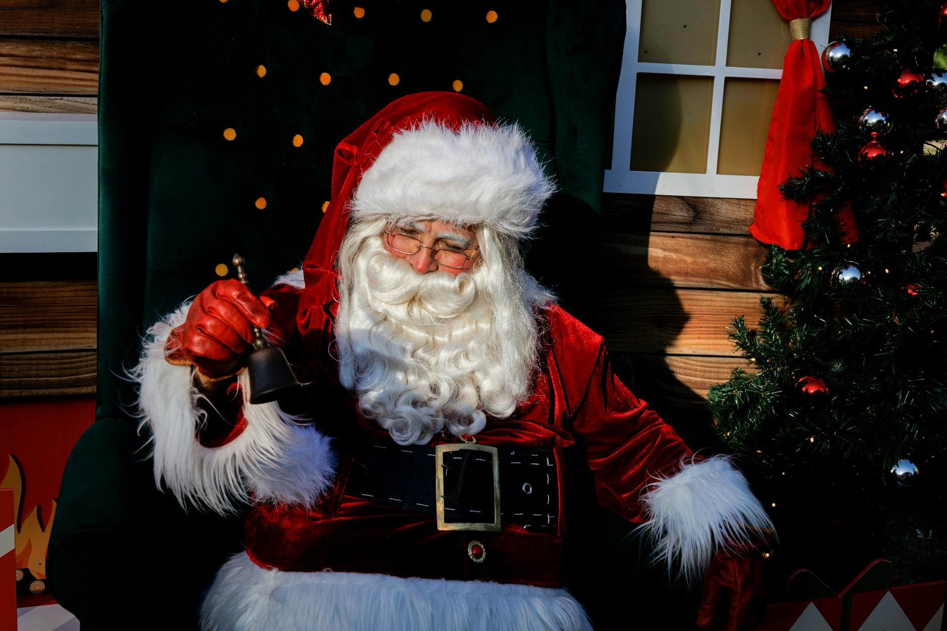 Festive Santa Claus ringing a bell next to a decorated Christmas tree indoors.