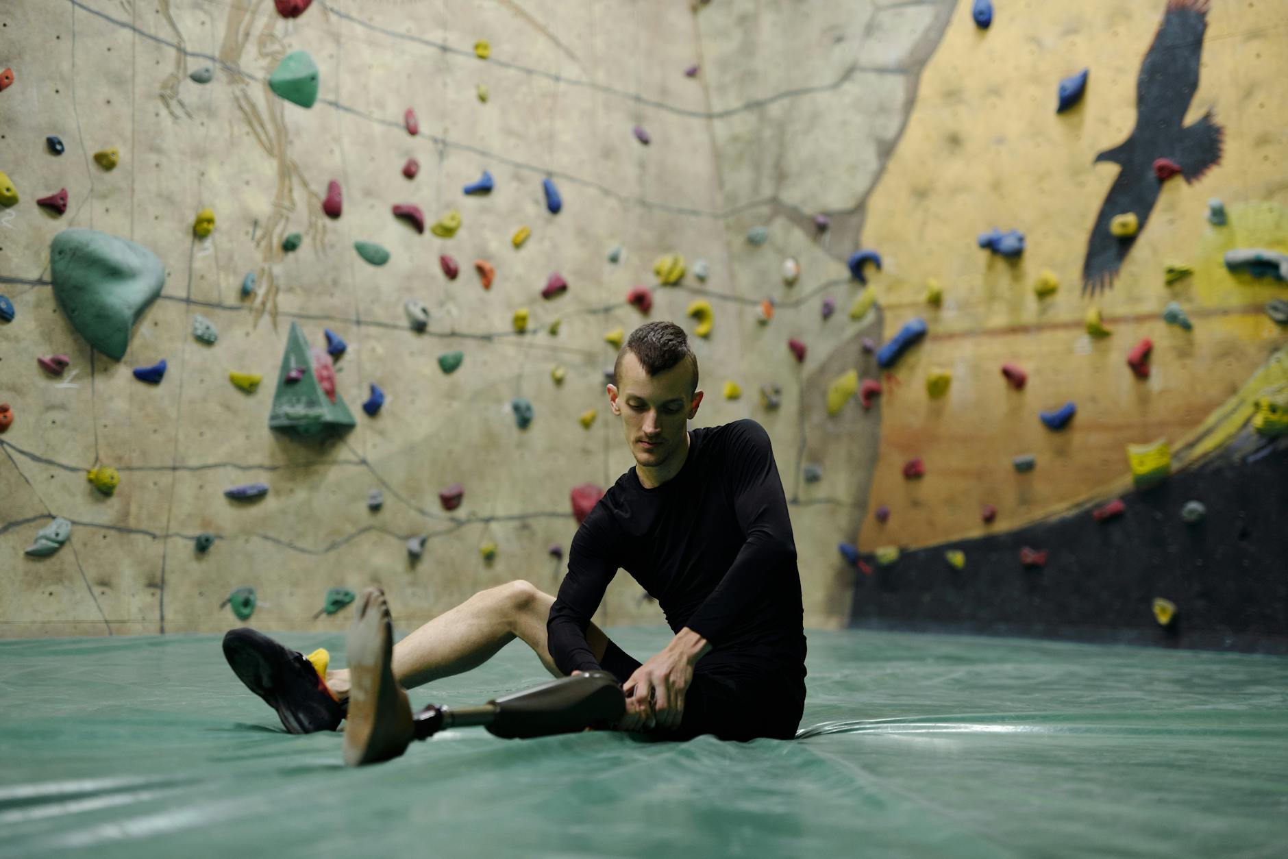 A male amputee with a prosthetic leg sits on the ground inside an indoor climbing gym.