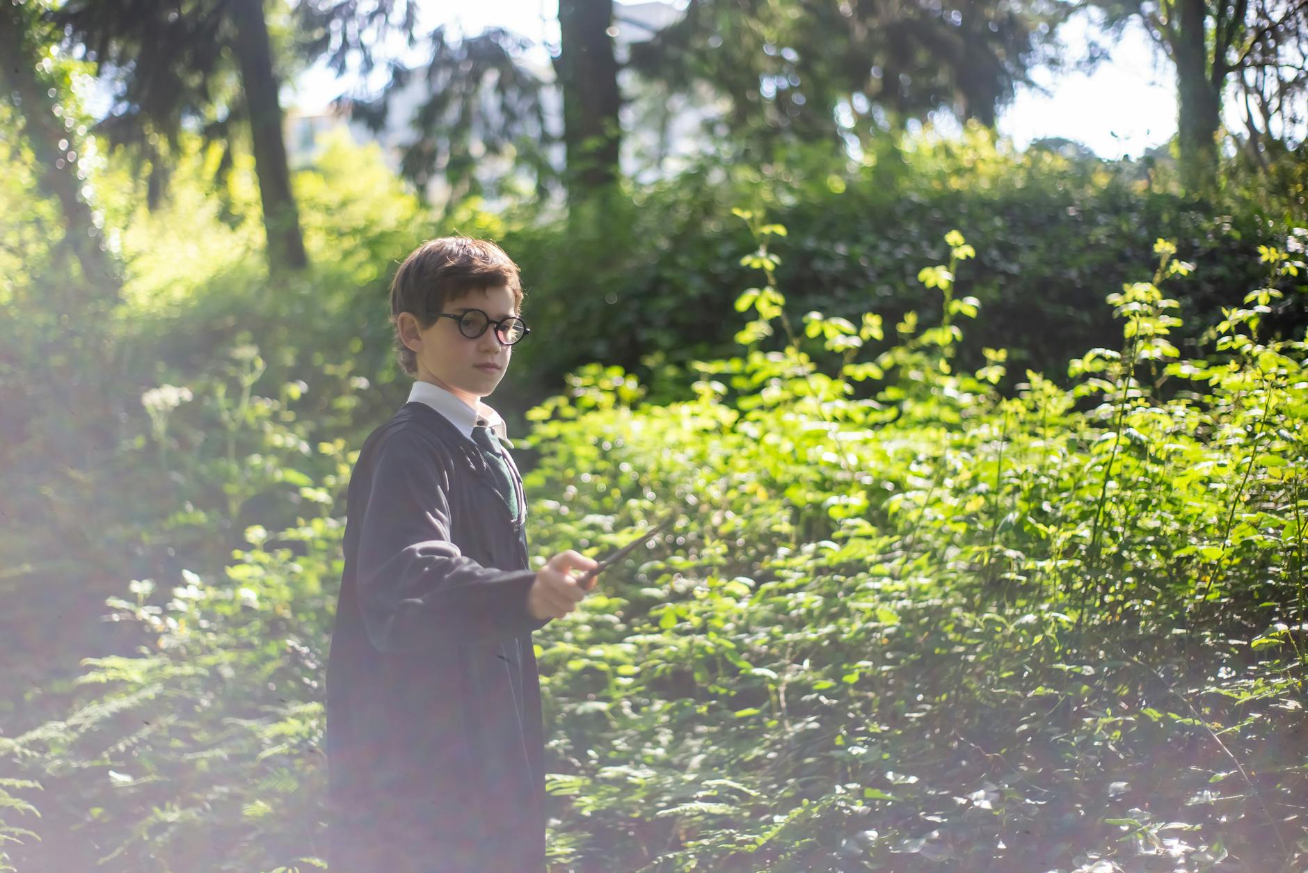A young boy in glasses role plays as a wizard with a wand in a lush green forest.