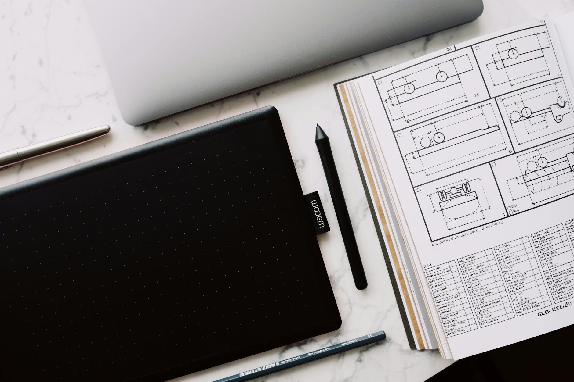 From above of black graphic tablet with marker and pen aside located in organized order on marble table near open book with drawn scheme